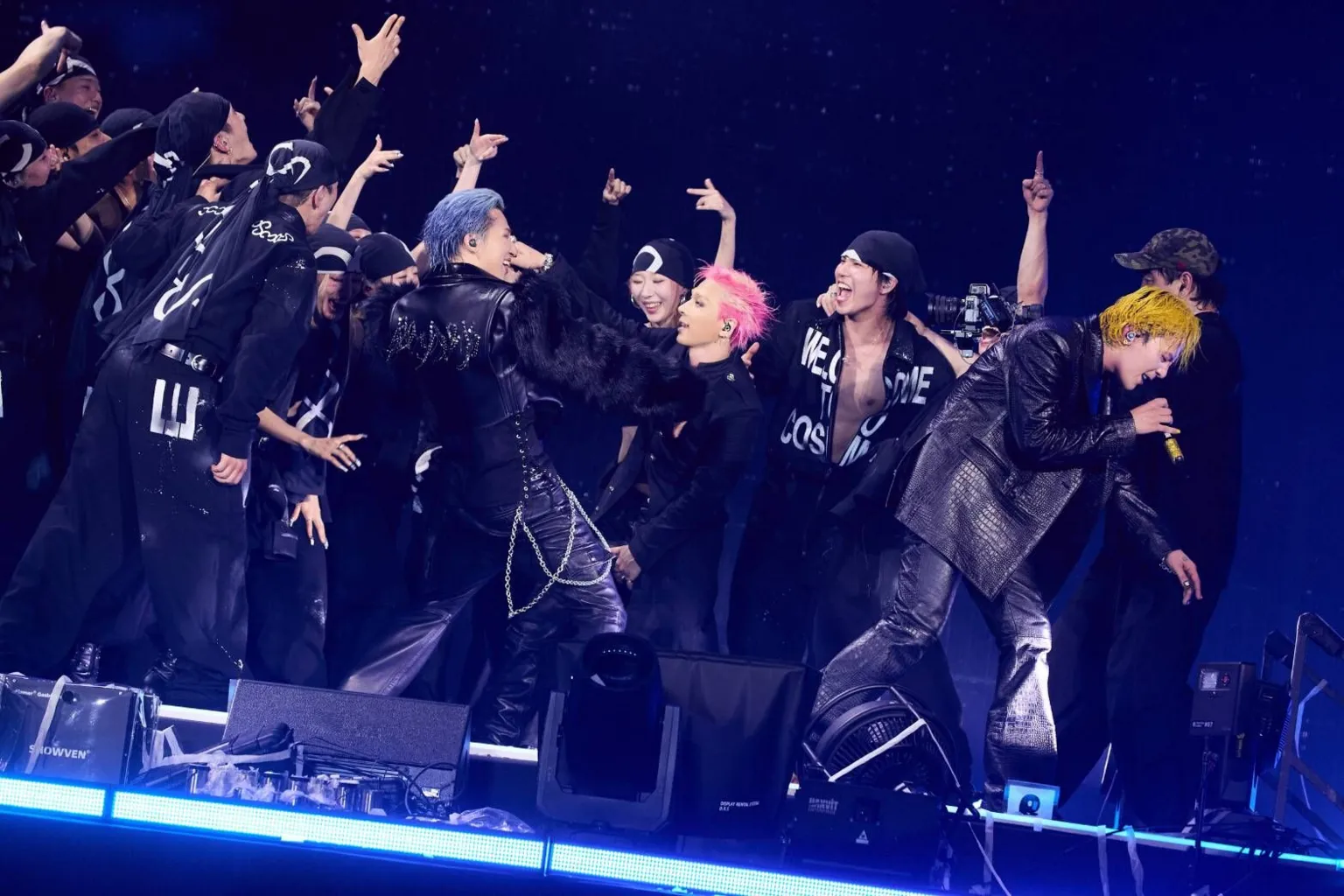 YG Entertainment A performer with bright yellow hair leans forward at the front of a stage, holding a handheld microphone. Around the performer, a group of performers and dancers in black outfits gather in a tight formation, with several raising their arms. Another performer with blue hair and a black outfit stands near the centre, while a performer with pink hair is visible beside them. They are illuminated by blue lighting against a dark background.