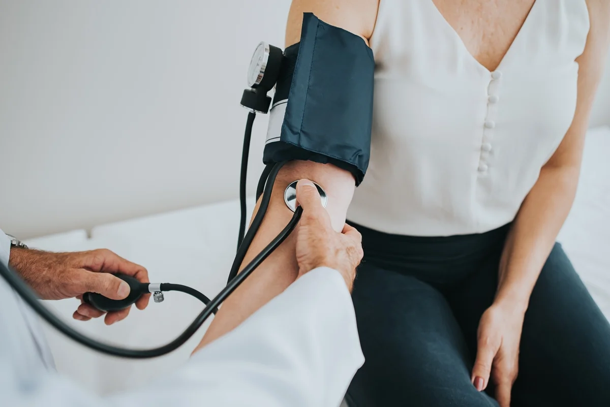 Imagem mostra mulher sentada em maca e tendo a pressão aferida com um aparelho manual - Metrópoles - pressão alta